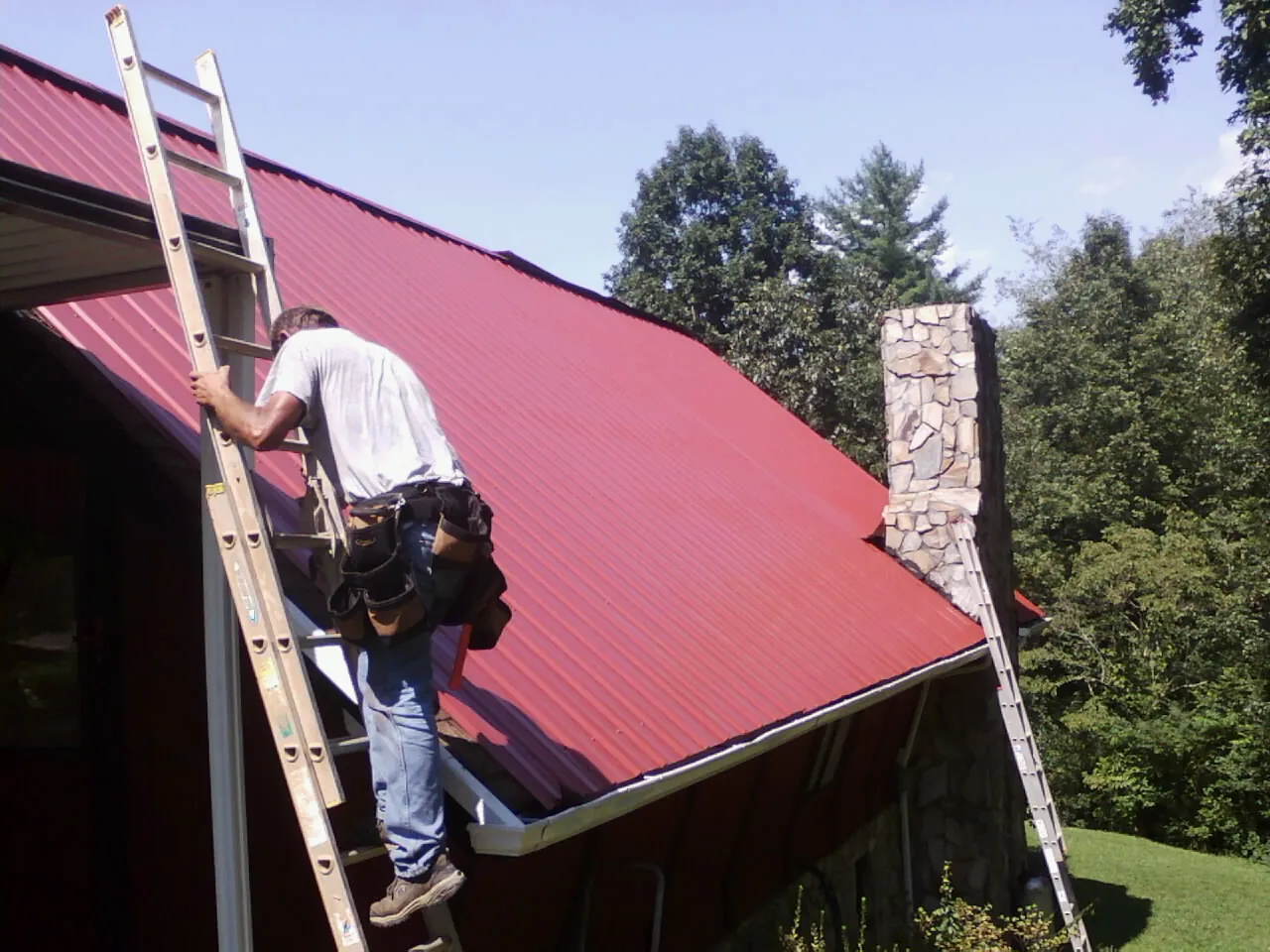 Roofer on a ladder beside a red metal roof and stone chimney — Soffit & Fascia Repair by Roofing Brothers in Nauvoo